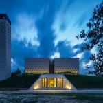 Night photograph of Aman Mosque with illuminated triangular perforations, a tall minaret and an earth-bermed entrance glowing from within