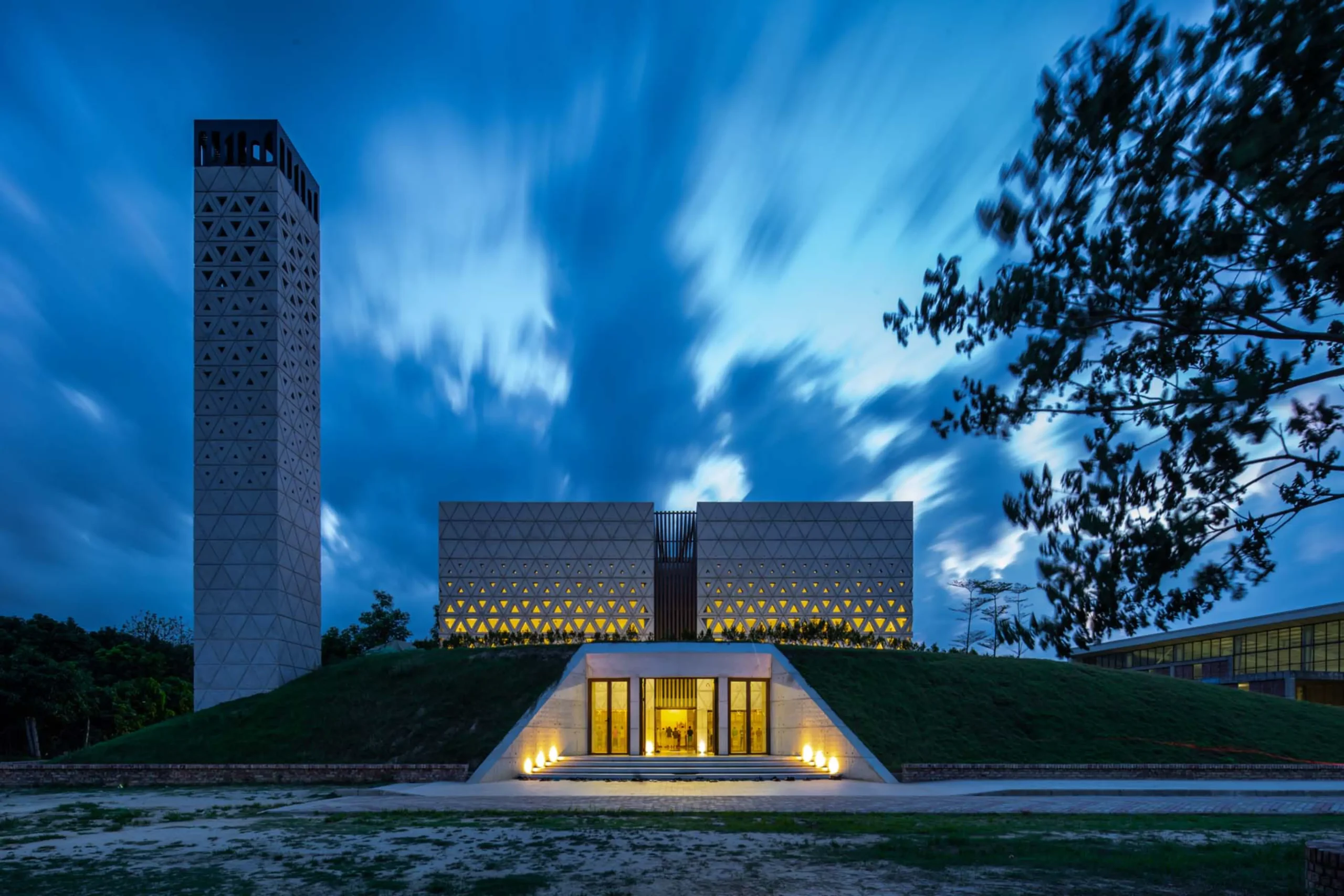 Nakshabid Architects_Night photograph of Aman Mosque with illuminated triangular perforations, a tall minaret and an earth-bermed entrance glowing from within