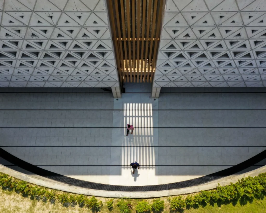 Overhead view of a sunlit courtyard between concrete walls patterned with triangular openings, with two people walking below