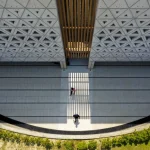 Overhead view of a sunlit courtyard between concrete walls patterned with triangular openings, with two people walking below