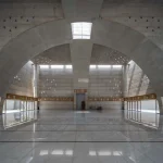 Interior of a large concrete prayer hall with intersecting arches, skylight and polished floor reflecting daylight