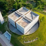 Aerial view of a cubic concrete mosque with triangular lattice façade and a tall perforated minaret set in green landscape