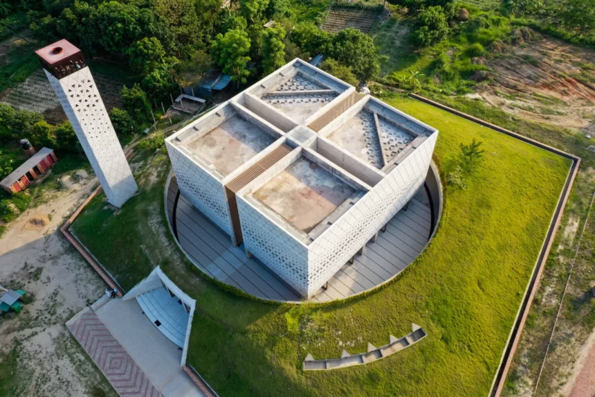 Aerial view of a cubic concrete mosque with triangular lattice façade and a tall perforated minaret set in green landscape