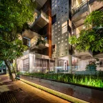 Night view of a multi-storey residential building with concrete and brick façade, glowing square openings, planted balconies and glass ground-floor spaces.
