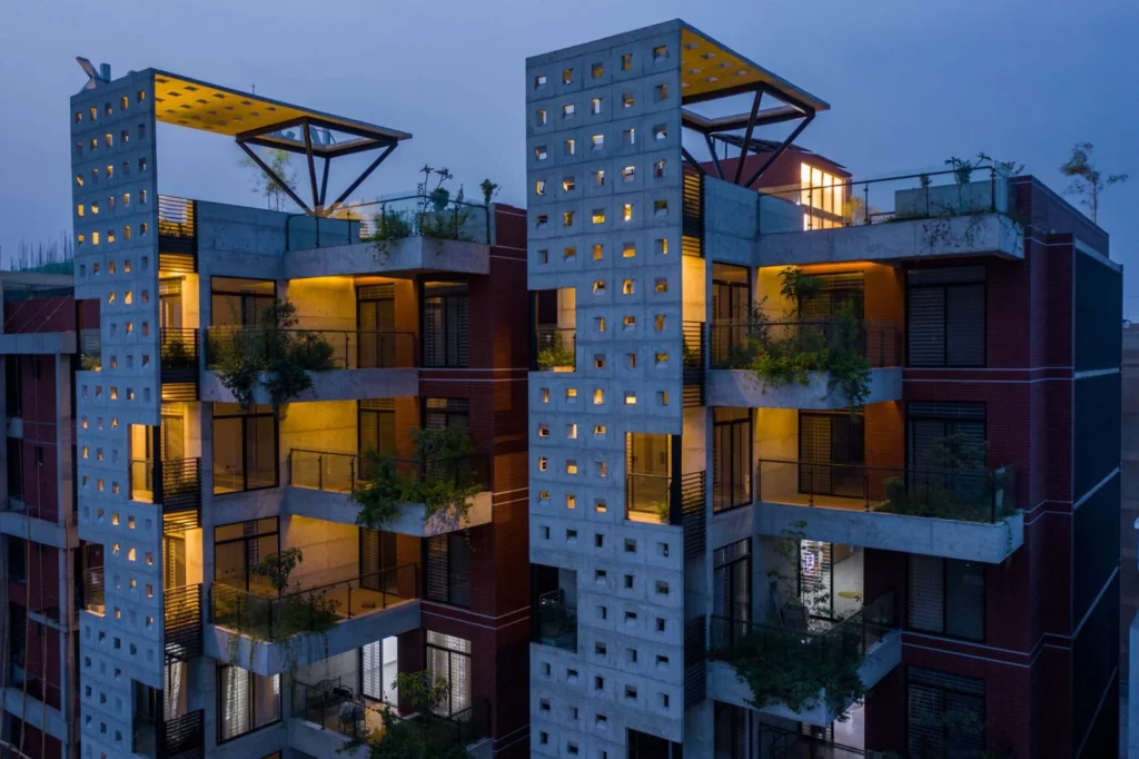 Night view of two concrete residential blocks with square window openings, planted balconies and warm interior lighting