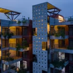 Night view of two concrete residential blocks with square window openings, planted balconies and warm interior lighting