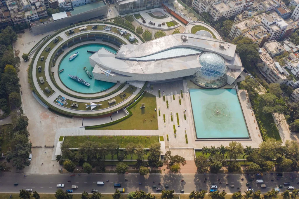 Aerial view of the Bangladesh Military Museum complex showing curved concrete forms, a large glass geodesic dome, water basins and outdoor military displays.