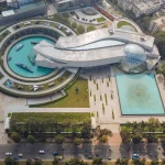 Aerial view of the Bangladesh Military Museum complex showing curved concrete forms, a large glass geodesic dome, water basins and outdoor military displays.