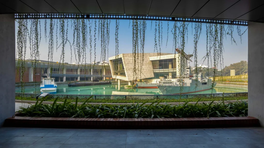 Framed view through hanging vines of naval vessels and a modern museum building beside a turquoise water channel