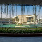 Framed view through hanging vines of naval vessels and a modern museum building beside a turquoise water channel