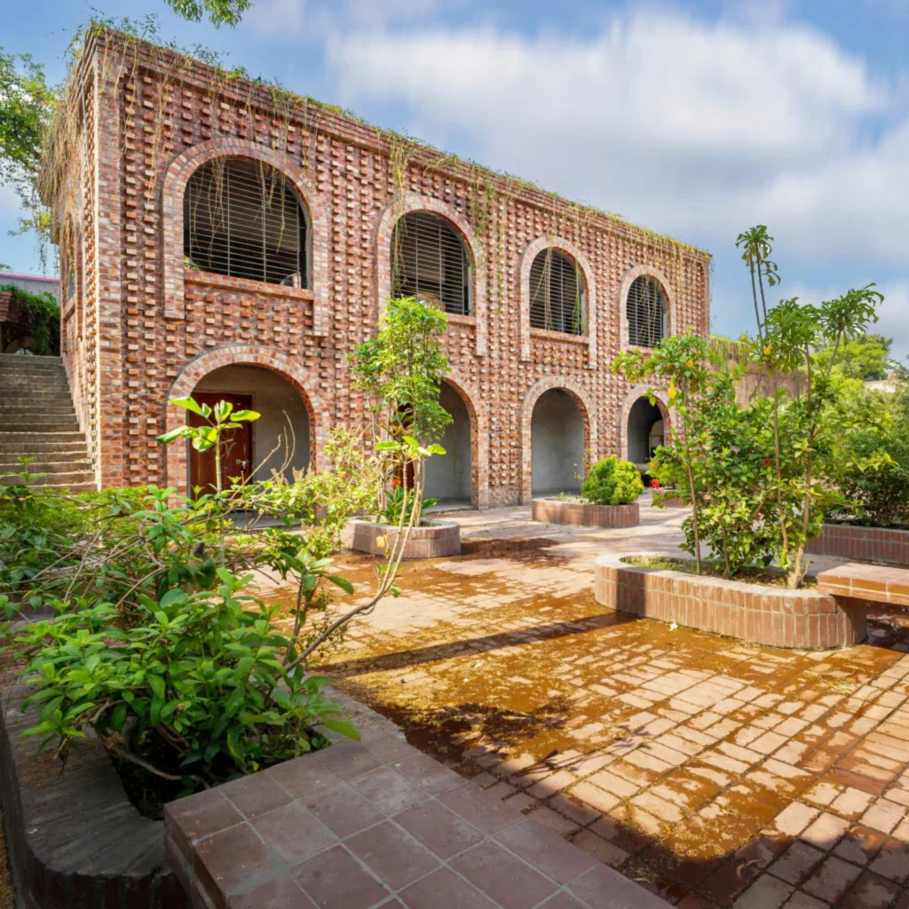 Two-storey red-brick building with arched openings and a planted courtyard in the foreground