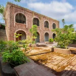 Two-storey red-brick building with arched openings and a planted courtyard in the foreground