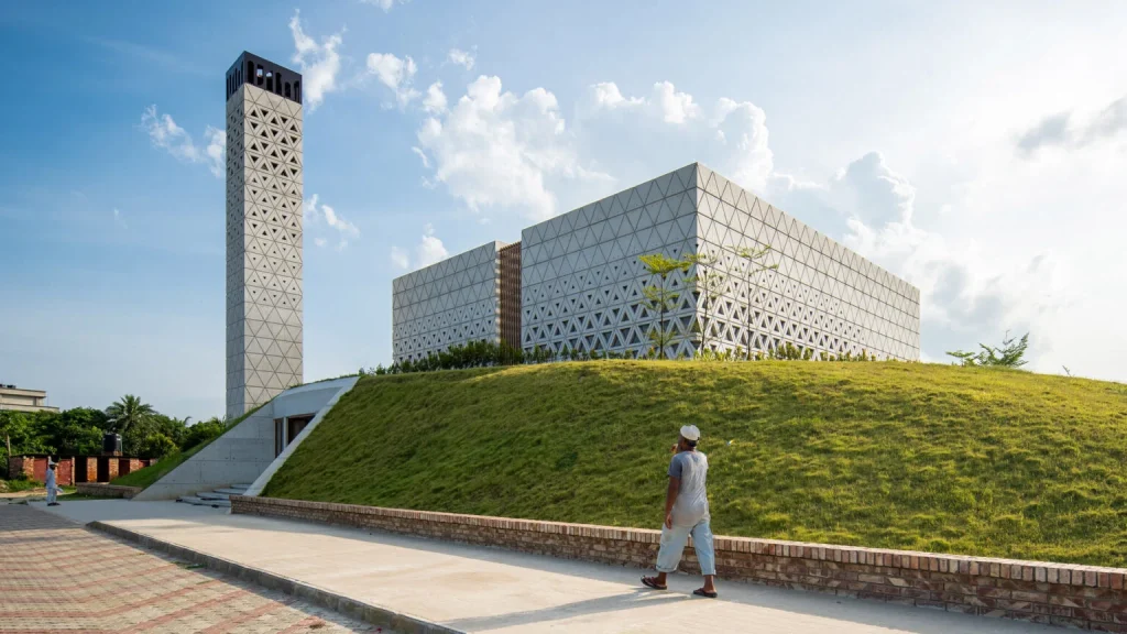 Man walking along a paved path toward a cubic concrete mosque with triangular lattice façade and a tall perforated minaret rising from a grassy berm