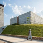 Man walking along a paved path toward a cubic concrete mosque with triangular lattice façade and a tall perforated minaret rising from a grassy berm