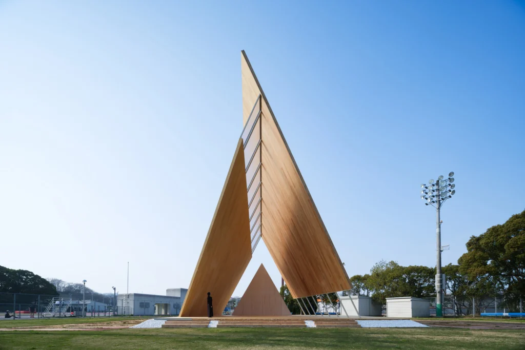 Onodatesho Pavilion_Front view of a tall, asymmetrical timber pavilion with a triangular canopy frame, featuring a smaller triangular timber podium inside and a person standing near the base against a clear blue sky.