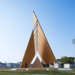 Front view of a tall, asymmetrical timber pavilion with a triangular canopy frame, featuring a smaller triangular timber podium inside and a person standing near the base against a clear blue sky.