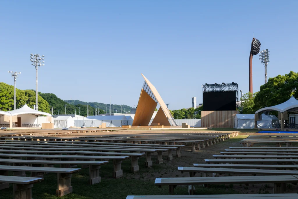 Wide view of an outdoor event site filled with rows of long wooden benches facing a tall triangular timber pavilion and large screen setups under a clear sky.