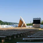 Wide view of an outdoor event site filled with rows of long wooden benches facing a tall triangular timber pavilion and large screen setups under a clear sky.
