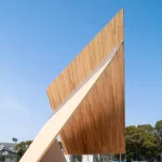 Dynamic side-angle view of the sharp, soaring timber pavilion structure displaying laminated wood paneling against a bright blue sky.