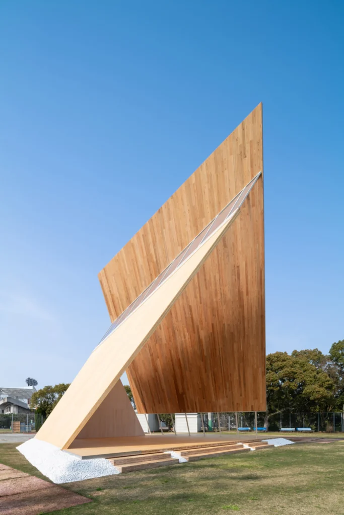 Dynamic side-angle view of the sharp, soaring timber pavilion structure displaying laminated wood paneling against a bright blue sky.