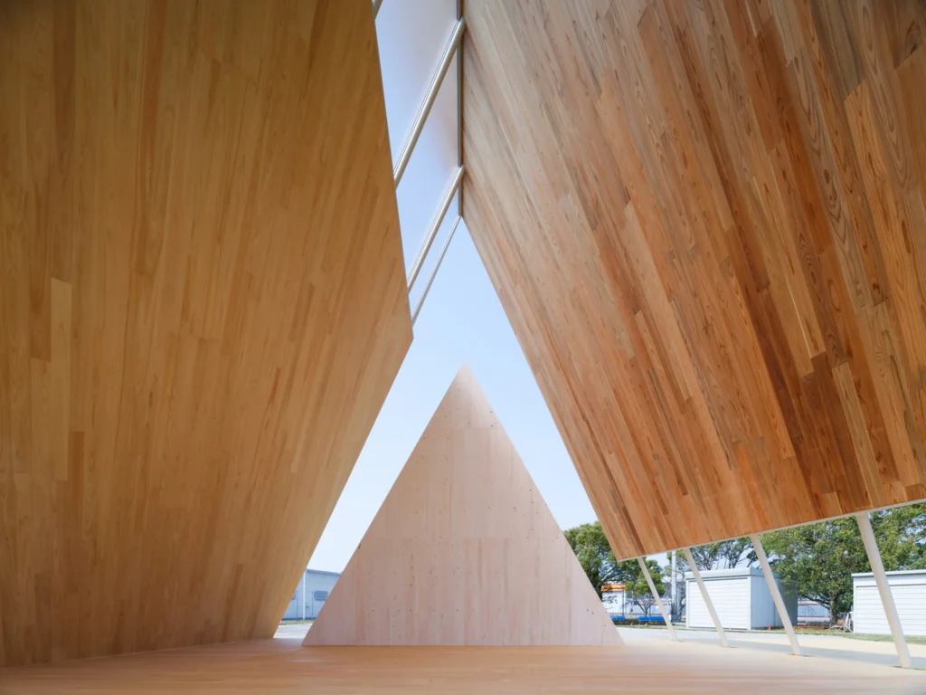 Interior view looking up beneath the angled timber roof panels toward a central triangular wooden element, framed by slanting support columns.