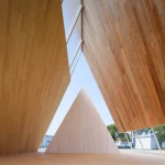 Interior view looking up beneath the angled timber roof panels toward a central triangular wooden element, framed by slanting support columns.