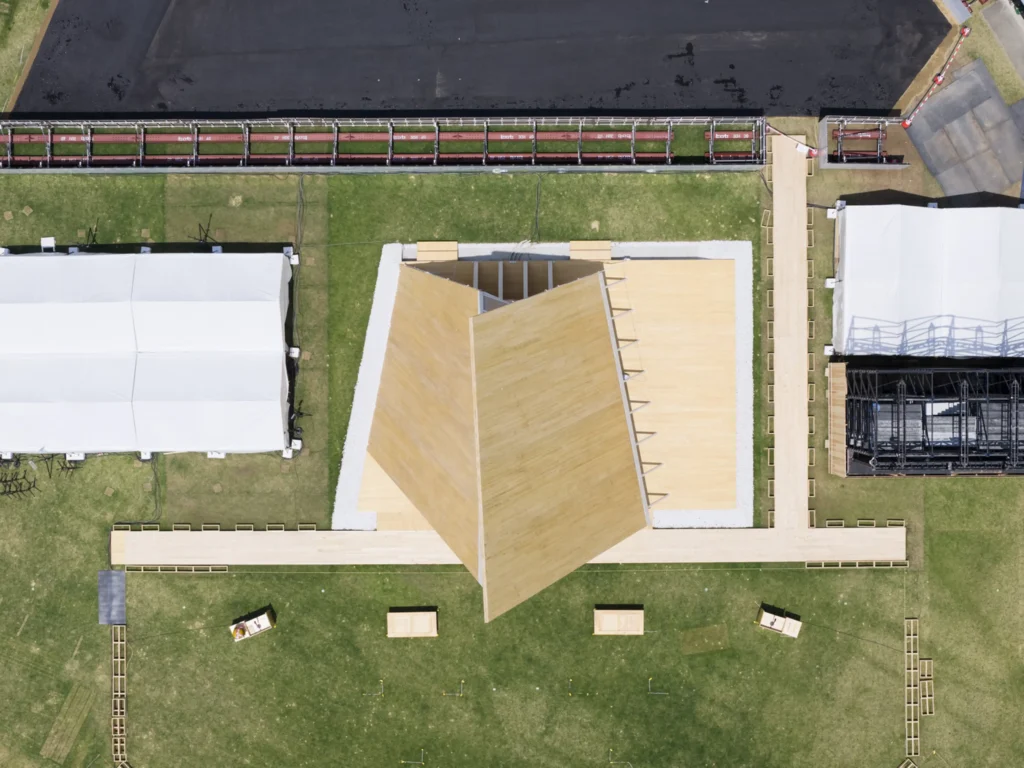 Direct overhead aerial drone view of the triangular timber pavilion, flanked by white temporary event tents and wooden walkways on a grassy field.