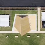 Direct overhead aerial drone view of the triangular timber pavilion, flanked by white temporary event tents and wooden walkways on a grassy field.