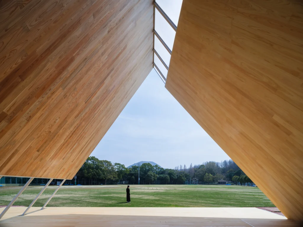 Interior view looking out from underneath the massive timber canopy frame toward an open green field, with a person standing in the distance framed by the structure.