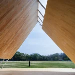Interior view looking out from underneath the massive timber canopy frame toward an open green field, with a person standing in the distance framed by the structure.