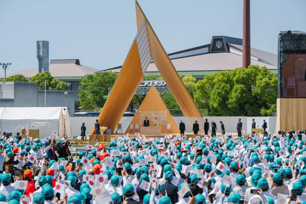 A large crowd holding small Japanese flags seated outdoors facing the triangular wooden stage during an official ceremony with dignitaries.