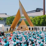A large crowd holding small Japanese flags seated outdoors facing the triangular wooden stage during an official ceremony with dignitaries.