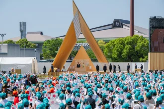 A large crowd holding small Japanese flags seated outdoors facing the triangular wooden stage during an official ceremony with dignitaries.