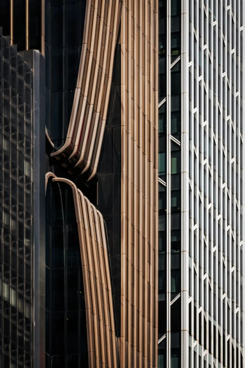 Architectural close-up of a building facade showing curved bronze vertical fins alongside a adjacent glass curtain wall with white structural elements.
