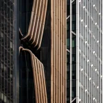 Architectural close-up of a building facade showing curved bronze vertical fins alongside a adjacent glass curtain wall with white structural elements.