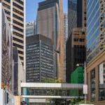 A bustling city street with pedestrians crossing the road, framed by an elevated pedestrian skybridge and modern office towers in the background.