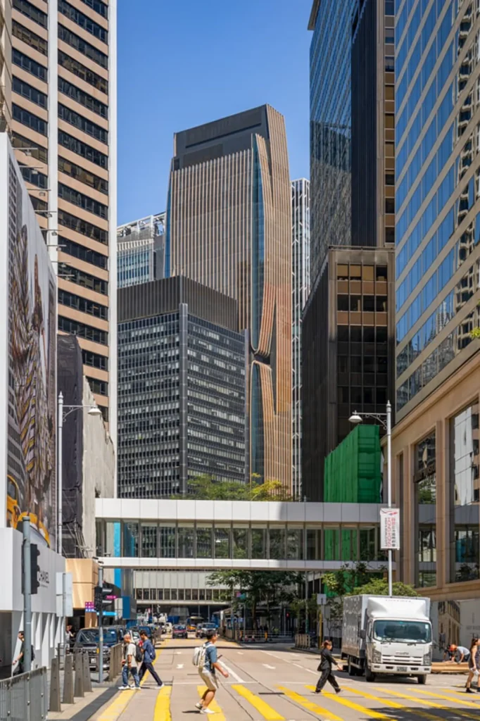 A bustling city street with pedestrians crossing the road, framed by an elevated pedestrian skybridge and modern office towers in the background.