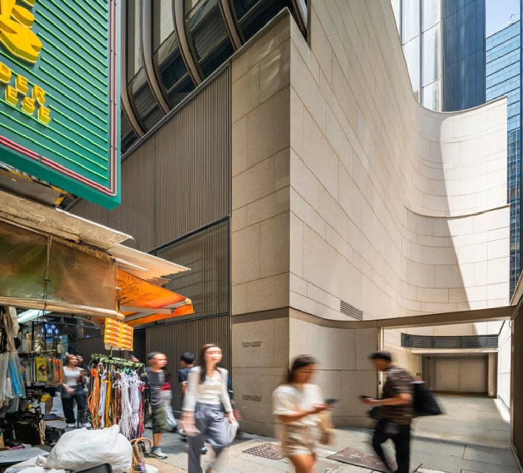 Pedestrians walking past a street-level market stall positioned next to a modern building with smooth, curved stone cladding.