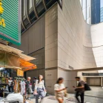 Pedestrians walking past a street-level market stall positioned next to a modern building with smooth, curved stone cladding.
