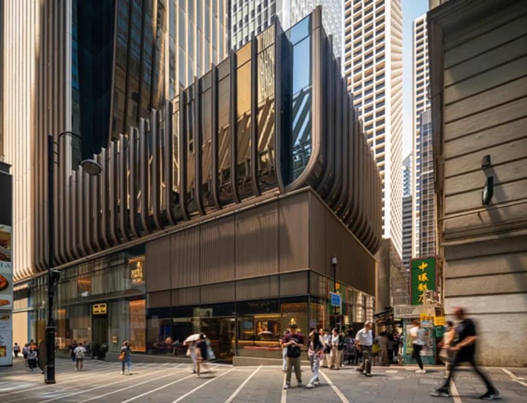 A low-angle exterior view of a multi-story building base wrapped in vertical bronze fins above ground-floor retail spaces and a pedestrian alleyway.