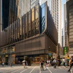 A low-angle exterior view of a multi-story building base wrapped in vertical bronze fins above ground-floor retail spaces and a pedestrian alleyway.