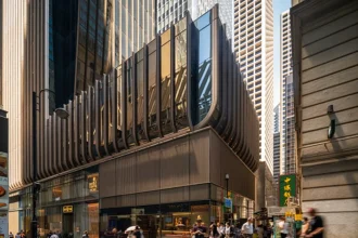 A low-angle exterior view of a multi-story building base wrapped in vertical bronze fins above ground-floor retail spaces and a pedestrian alleyway.