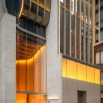 Night exterior view of a grand building entrance with glowing warm orange interior lighting visible through tall glass panels and stone pillars.