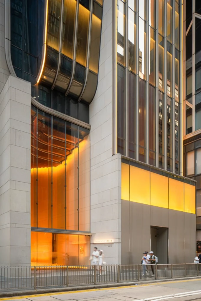 Night exterior view of a grand building entrance with glowing warm orange interior lighting visible through tall glass panels and stone pillars.
