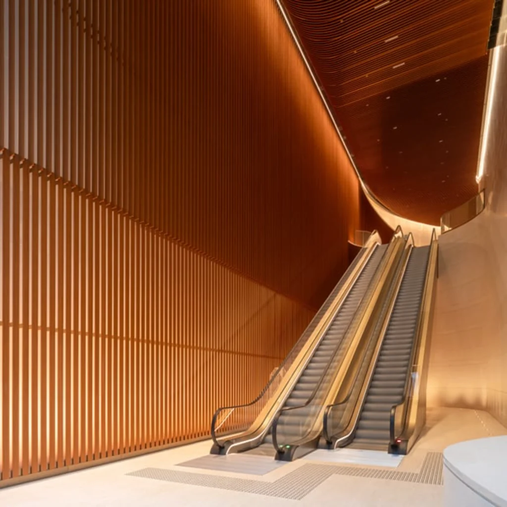 A modern interior lobby featuring parallel escalators set against a tall wall with vertical timber slats and a curving wooden slat ceiling with warm lighting.