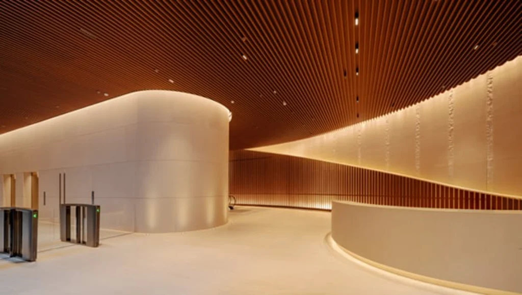 A sleek, minimalist building concourse with security turnstiles, smooth curved stone walls, and a continuous slatted wooden ceiling with cove lighting.