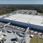 Aerial view of a large data center facility surrounded by forest, featuring industrial cooling systems, extensive piping, and electrical infrastructure
