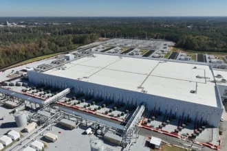Aerial view of a large data center facility surrounded by forest, featuring industrial cooling systems, extensive piping, and electrical infrastructure
