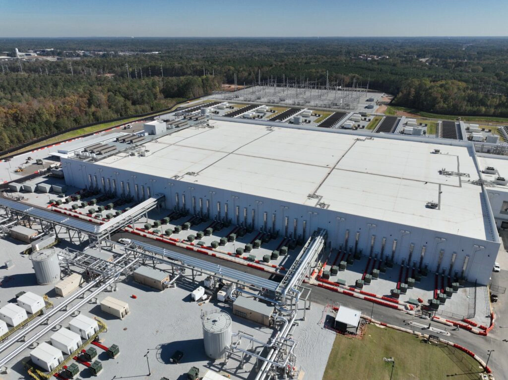 Aerial view of a large data center facility surrounded by forest, featuring industrial cooling systems, extensive piping, and electrical infrastructure