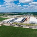 Aerial view of a large data center campus under construction, featuring multiple industrial buildings, electrical substations, and surrounding agricultural land
